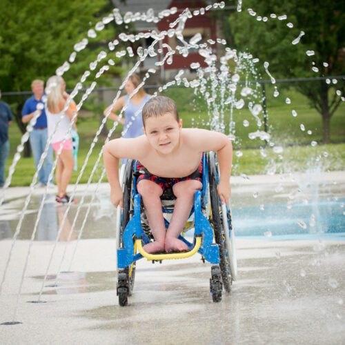 Child using a wheelchair moving through water jets at Parkersburg City Park