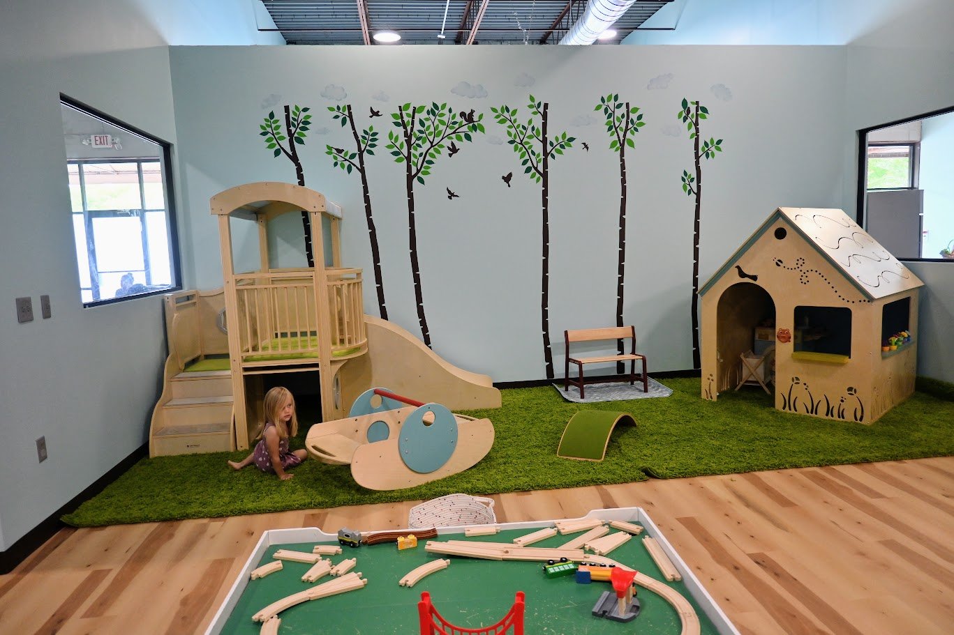 Forest-themed toddler corner with a small wooden slide, playhouse, and train table.