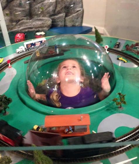 Child inside a clear dome on a train-table style exhibit at the museum.