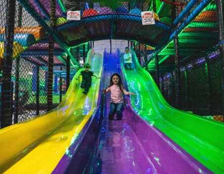 Children sliding down side-by-side slides inside the Rebounderz indoor playground.