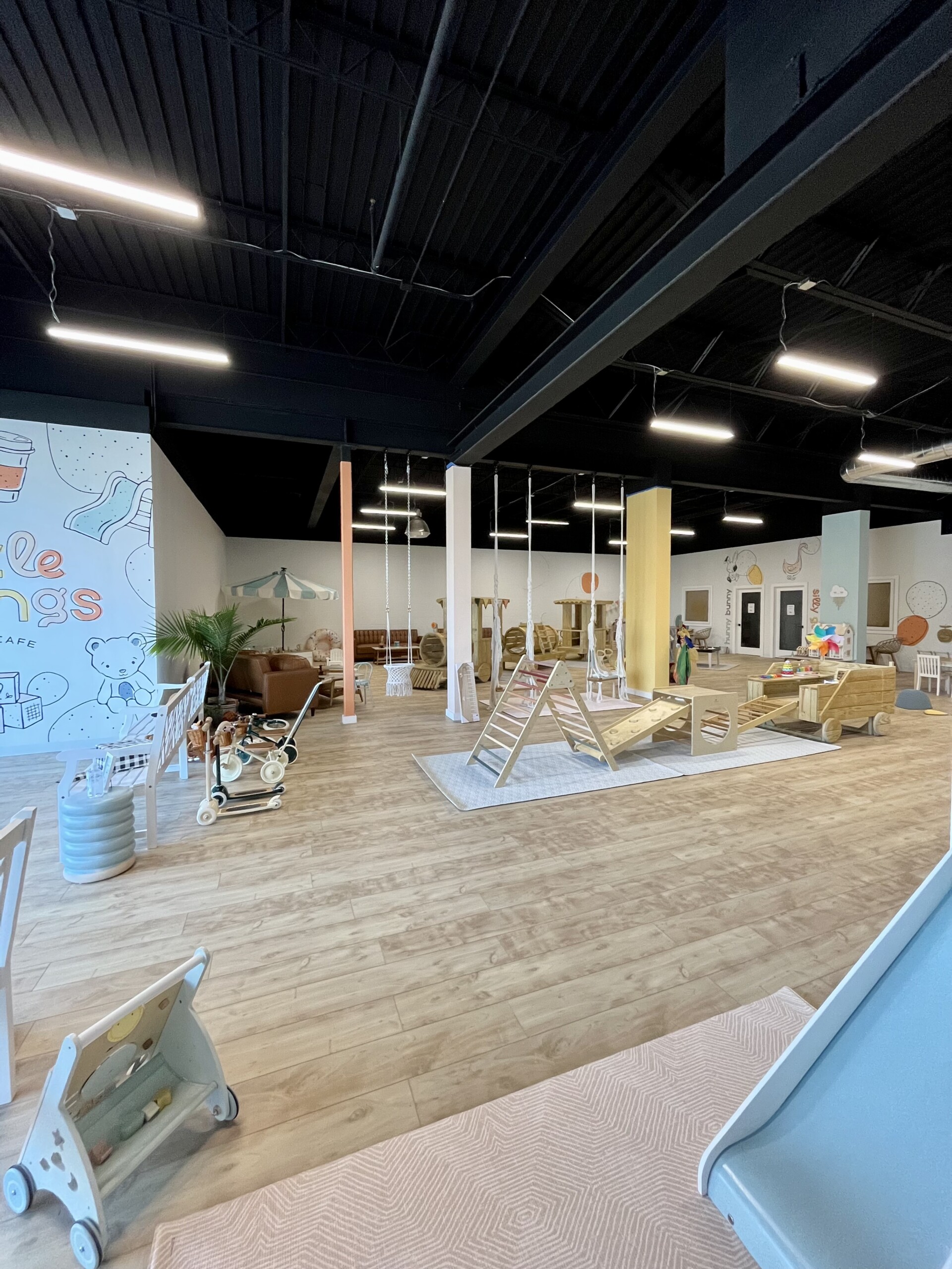 Wide view of The Little Things Play Cafe indoor play area with wooden climbing structures and swings.