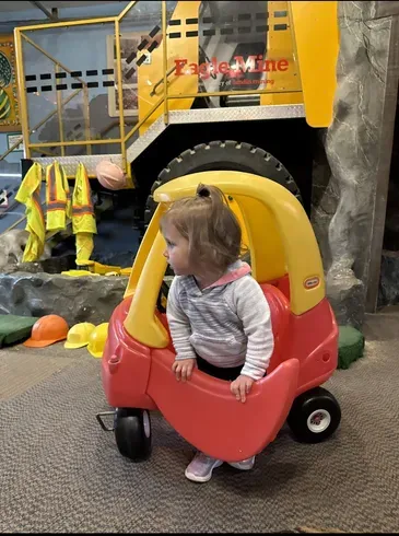 Child in a small ride-on car in front of a mining-themed museum exhibit.