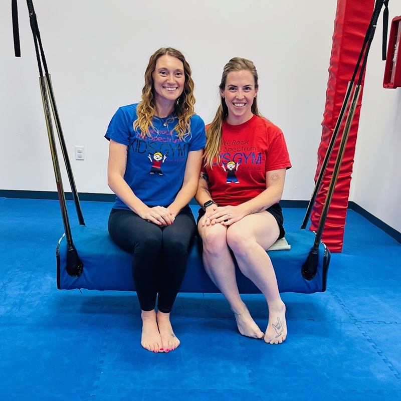 Two staff members on a large platform swing inside We Rock the Spectrum Kid's Gym in Jacksonville.