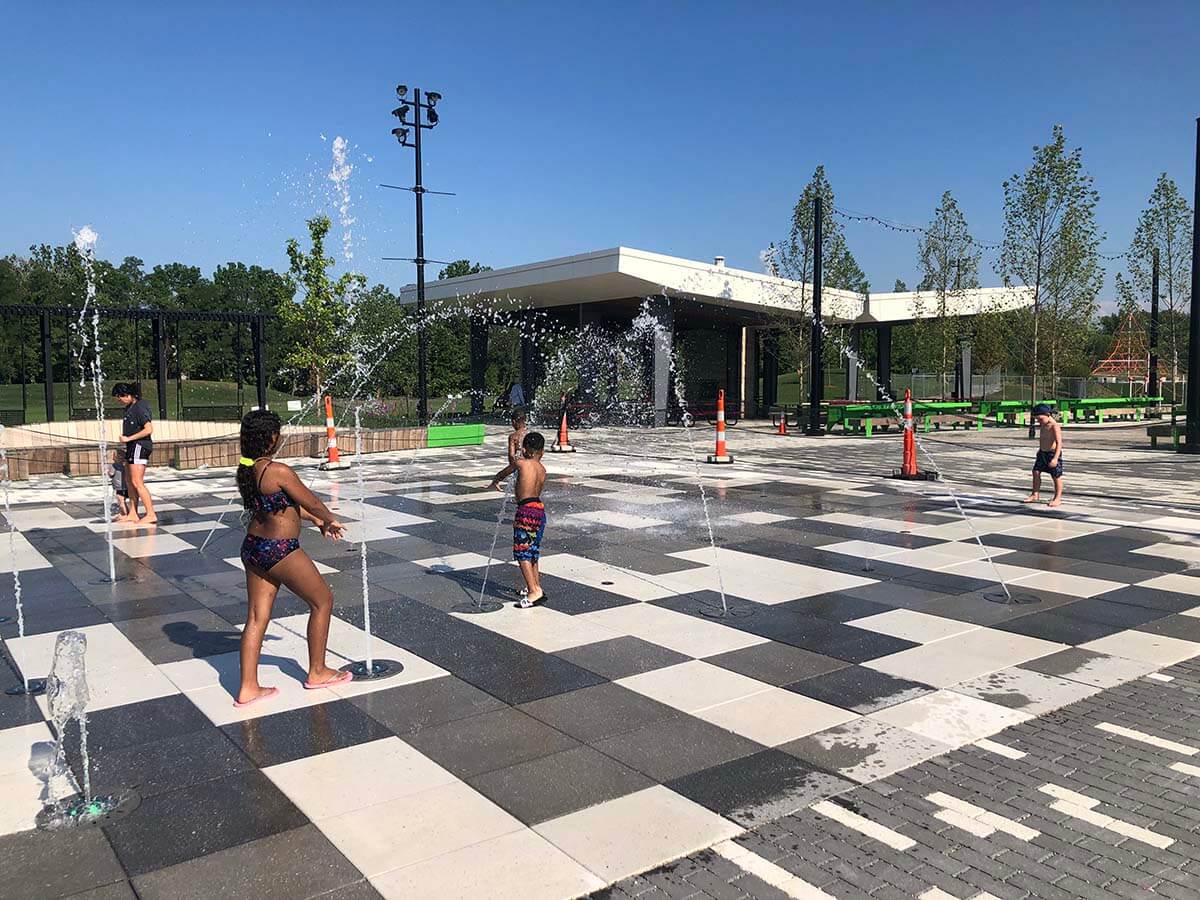 Kids play as water splashes from the Switchyard Plaza Spray Pad on a sunny day.